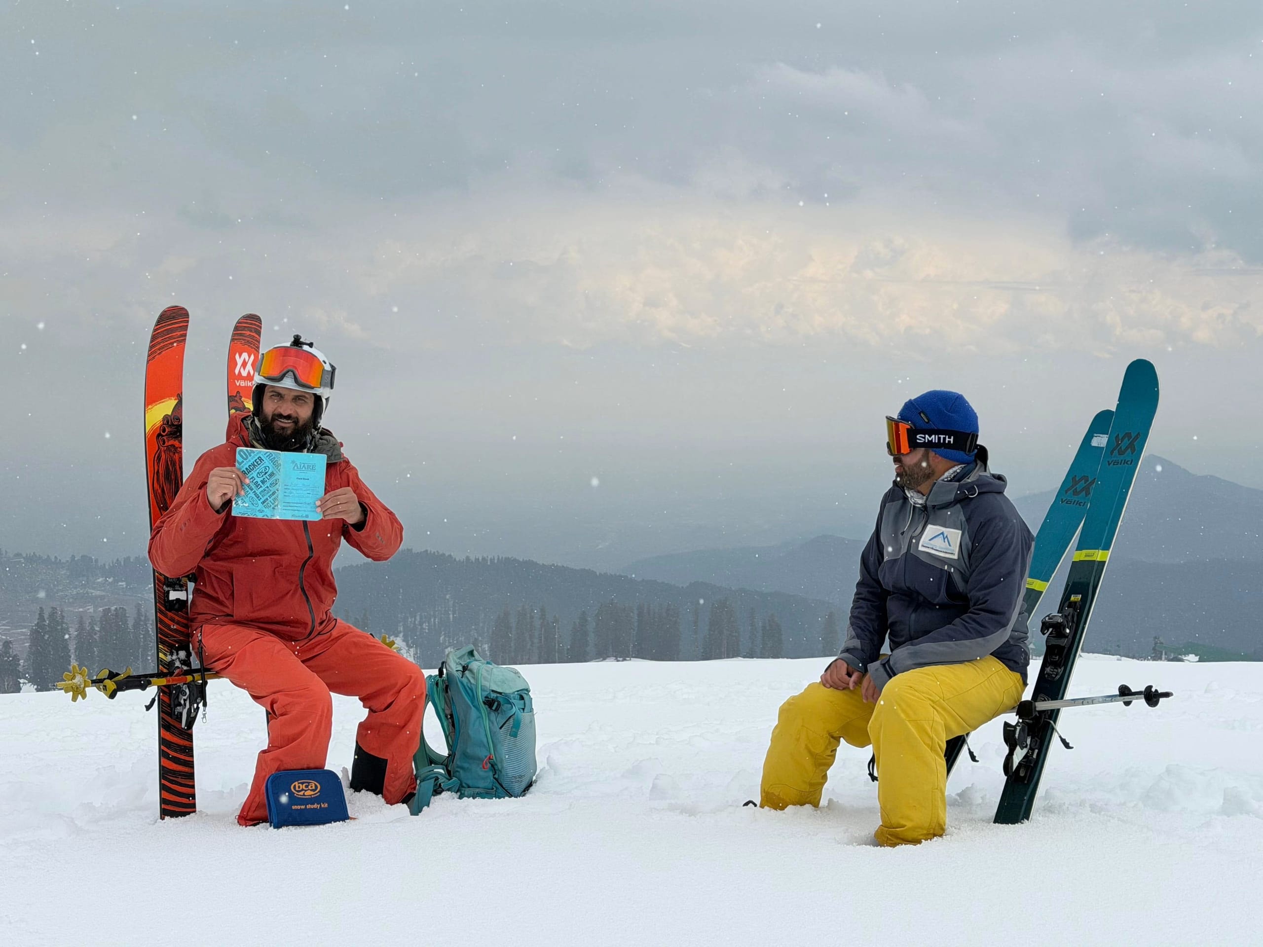 Two guides sitting on the snow with their skis planted beside them, one holding a snow study card, the valley behind.