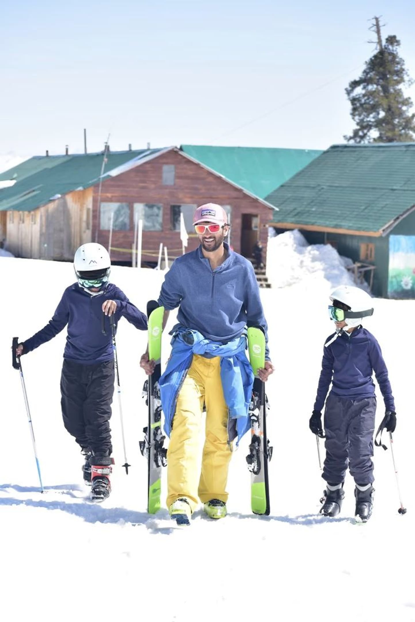 An adult and two children in helmets walking with their skis past traditional wooden buildings in Gulmarg.