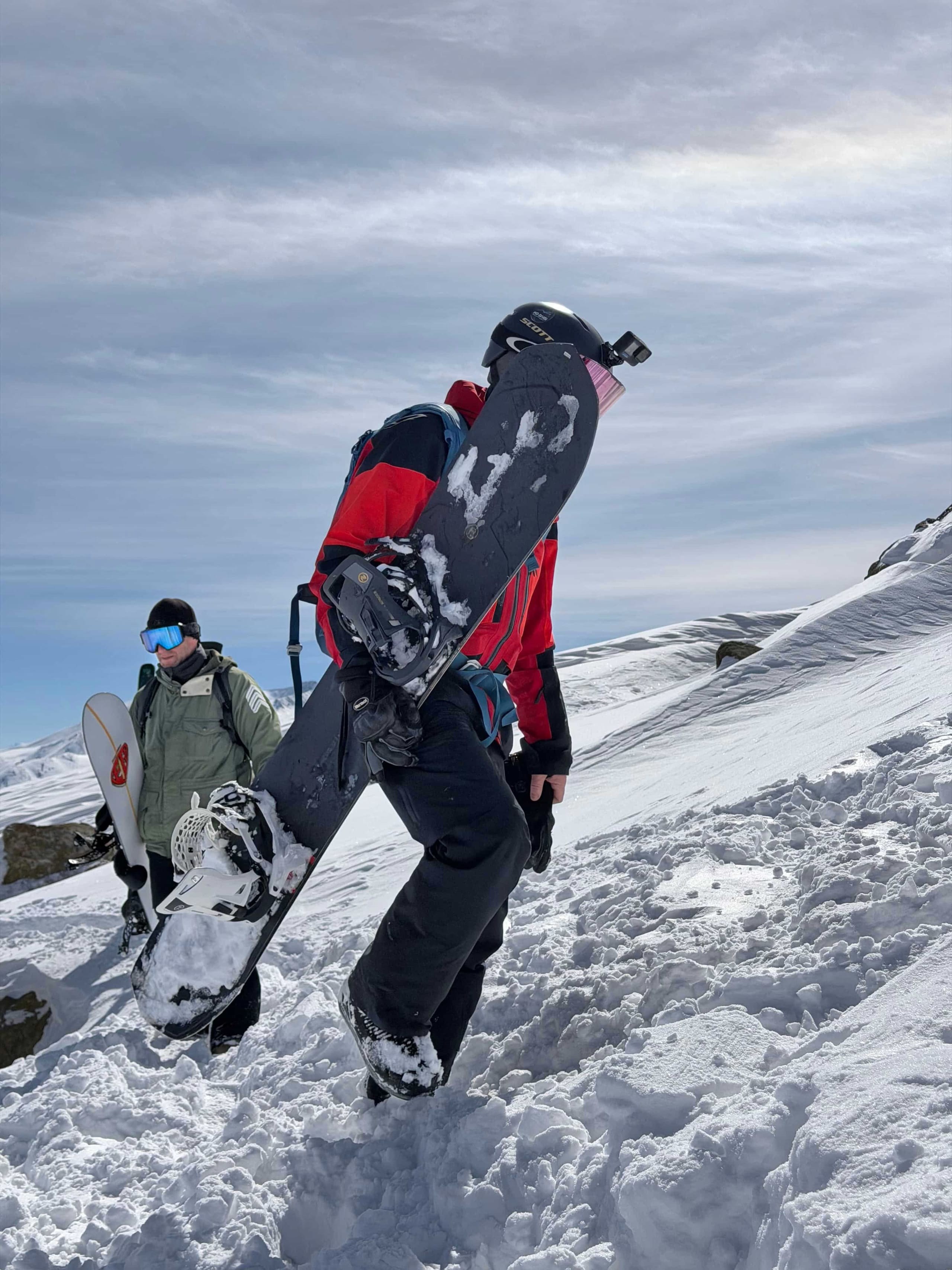 Two snowboarders carrying their boards up a snow covered ridge under a bright sky.