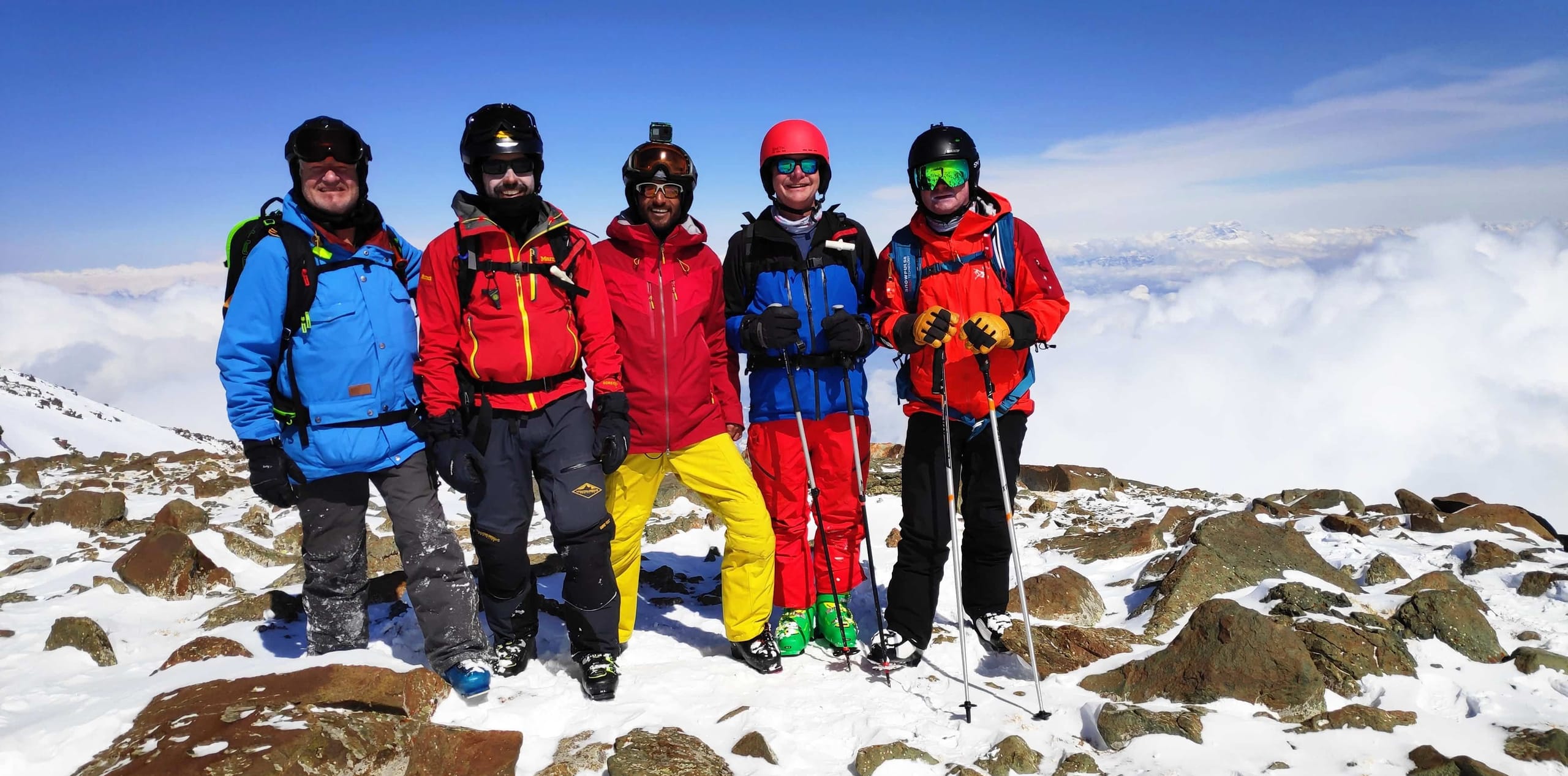 Five skiers standing together on a rocky snow covered summit above a sea of cloud.