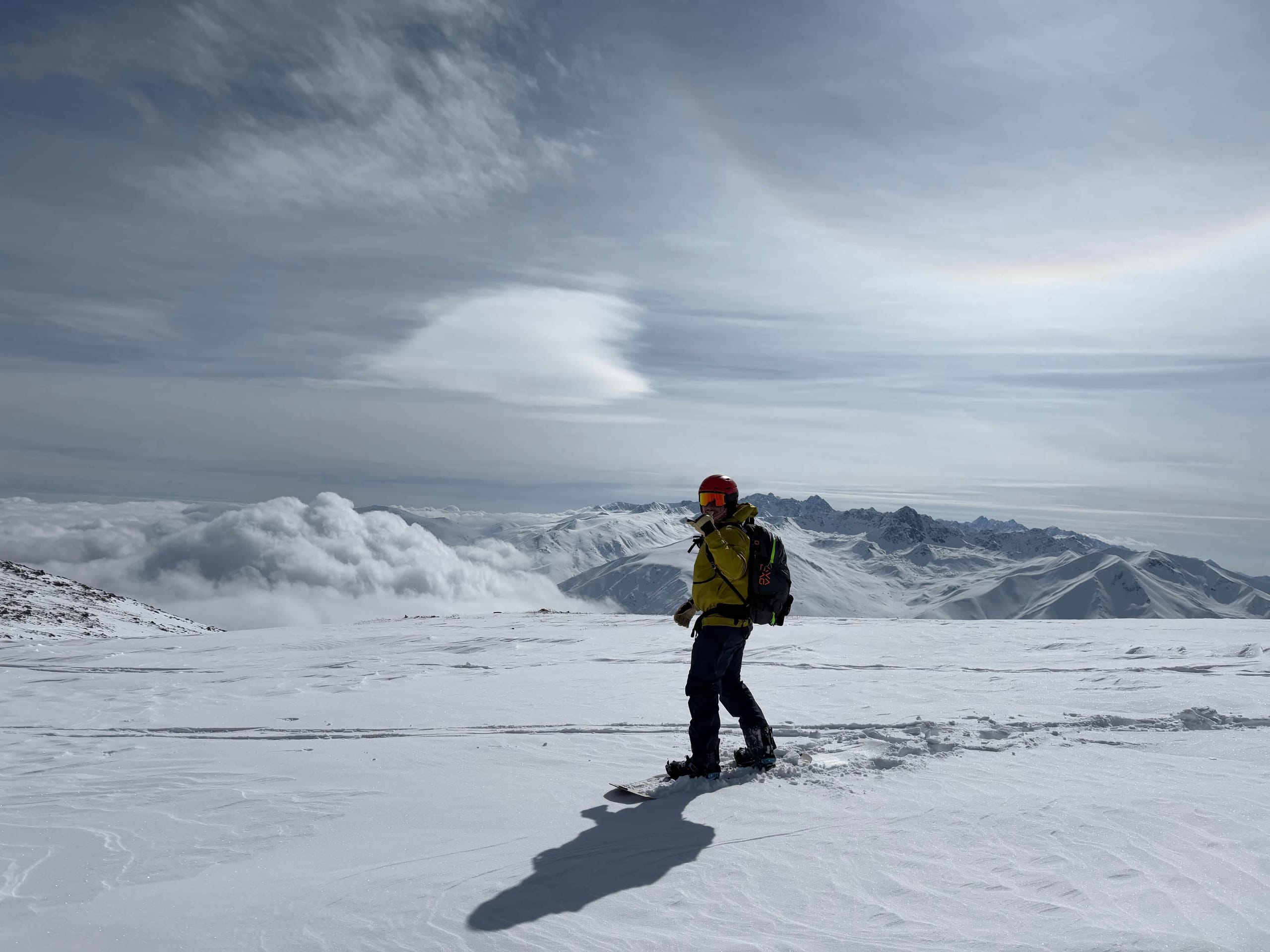 A snowboarder in a yellow jacket and red helmet standing on a wide open snowfield high above a sea of cloud, with mountain ranges on the horizon.