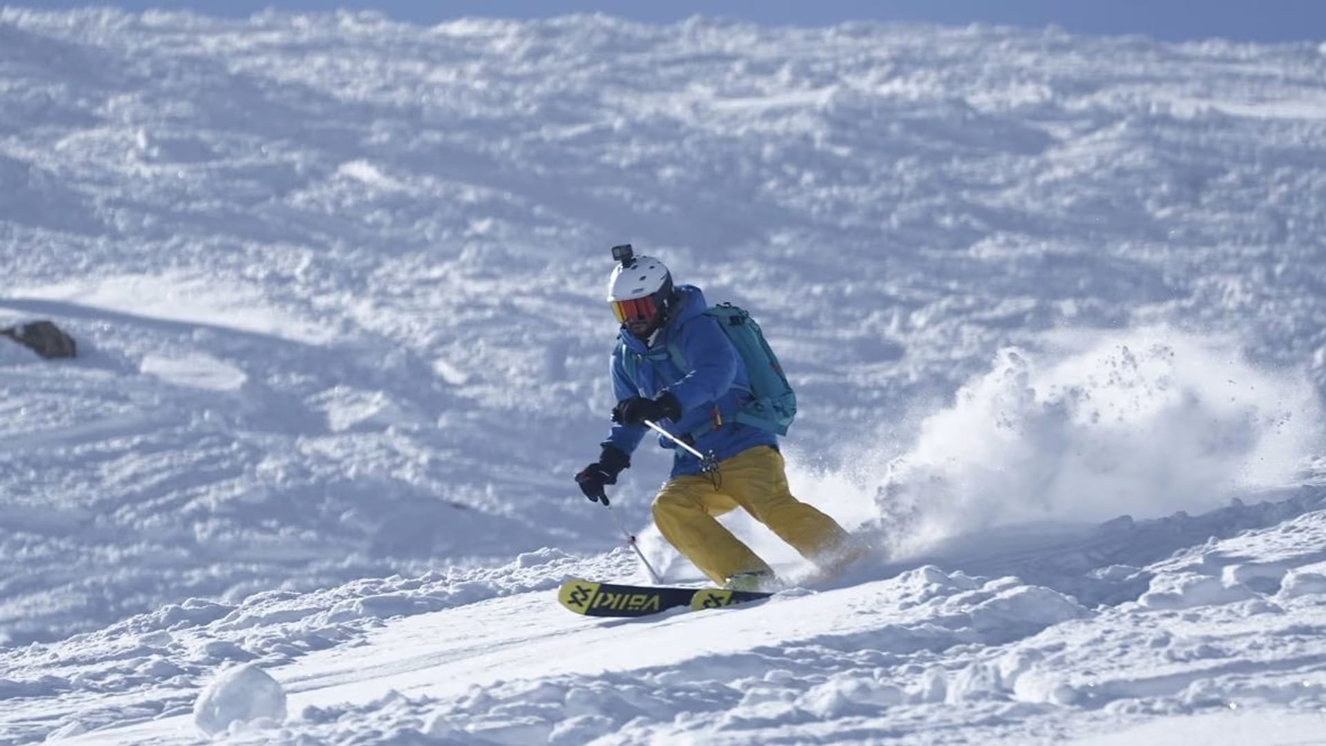 A snowboarder in a blue jacket and yellow trousers carving through chopped powder, throwing up a wall of spray.