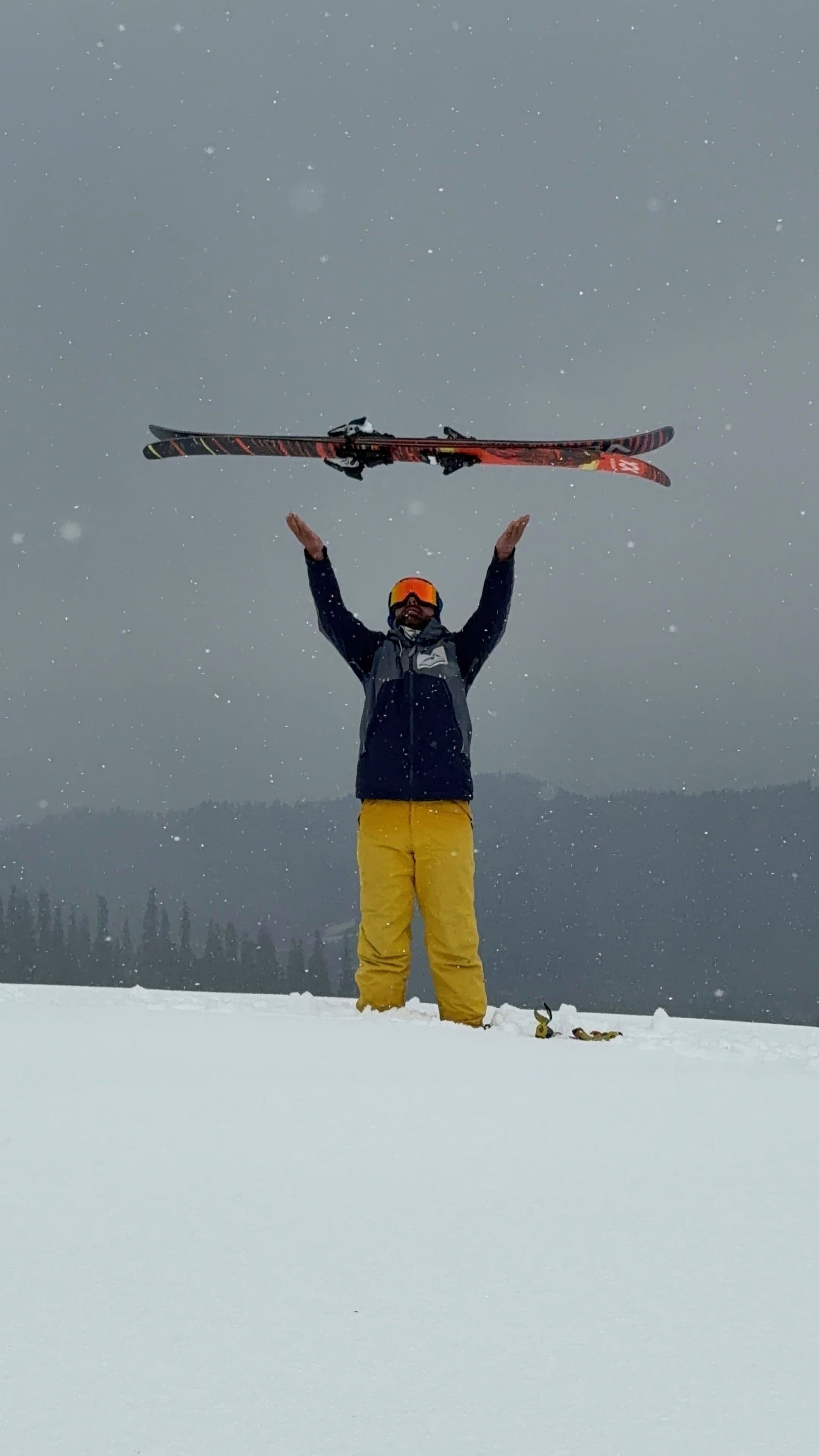 A skier holding their skis overhead with both arms raised as snow falls around them.