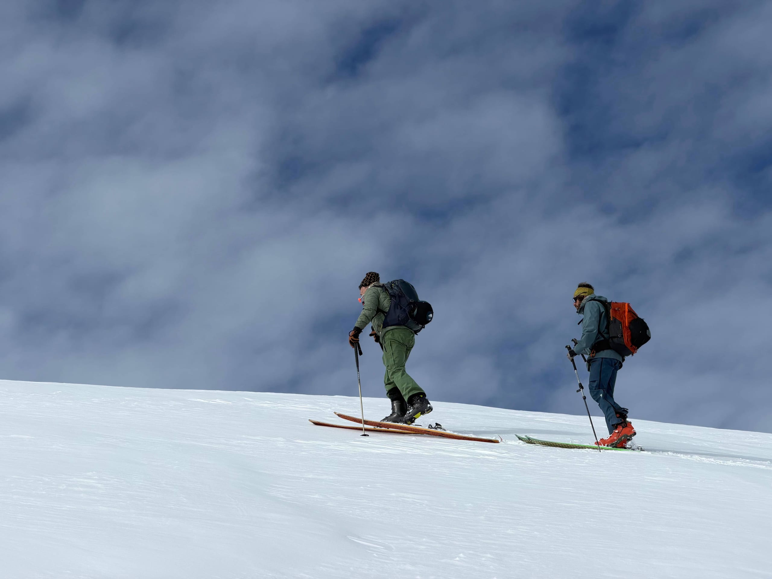 Two ski tourers skinning uphill in single file across a snow slope, with thick cloud filling the valley behind them.
