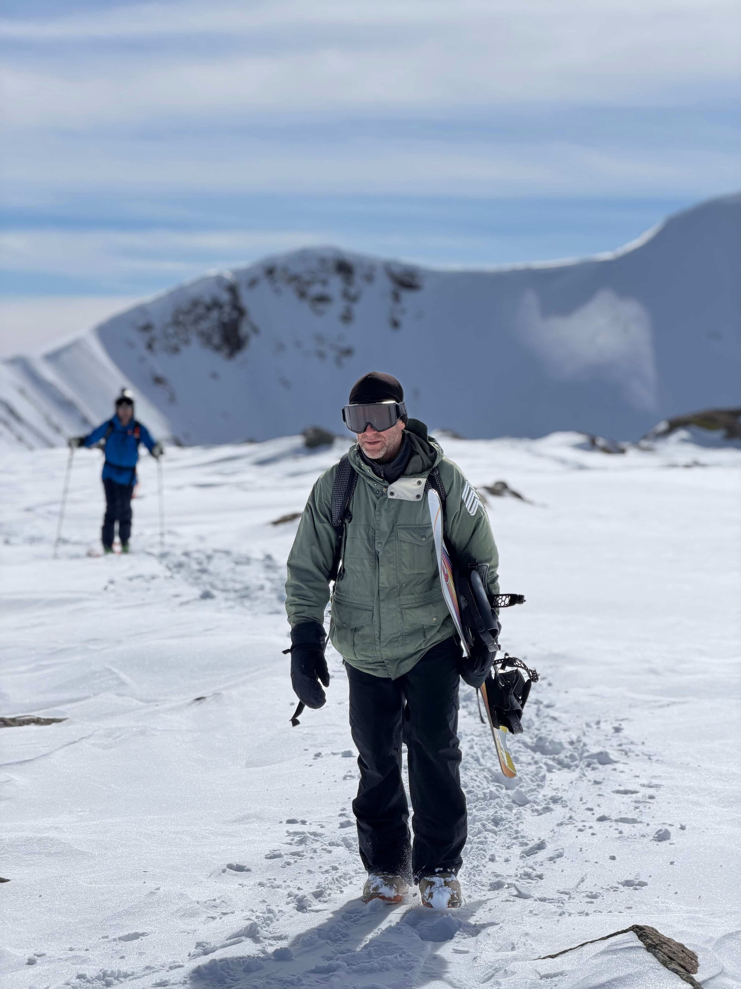A snowboarder walking down a snow slope carrying their board, with a second rider skinning up behind.