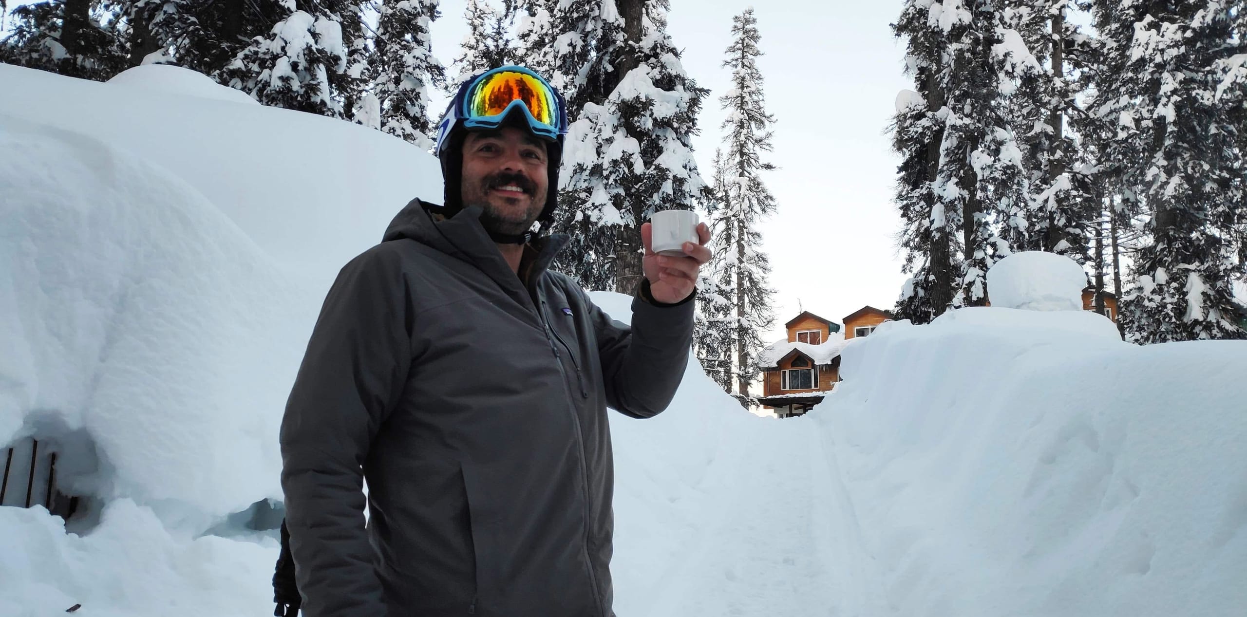 A man holding a phone beside deep snowbanks and snow laden pine trees in Gulmarg village.