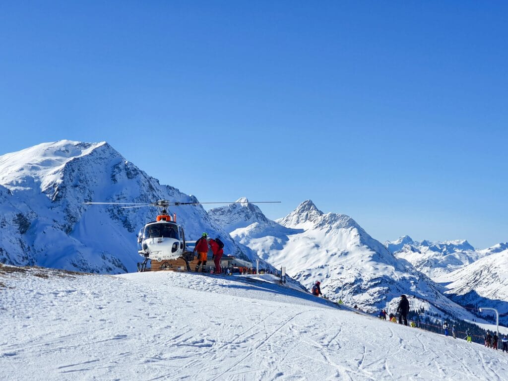 A helicopter rests on a snowy mountain with skiers and scenic Alpine views.